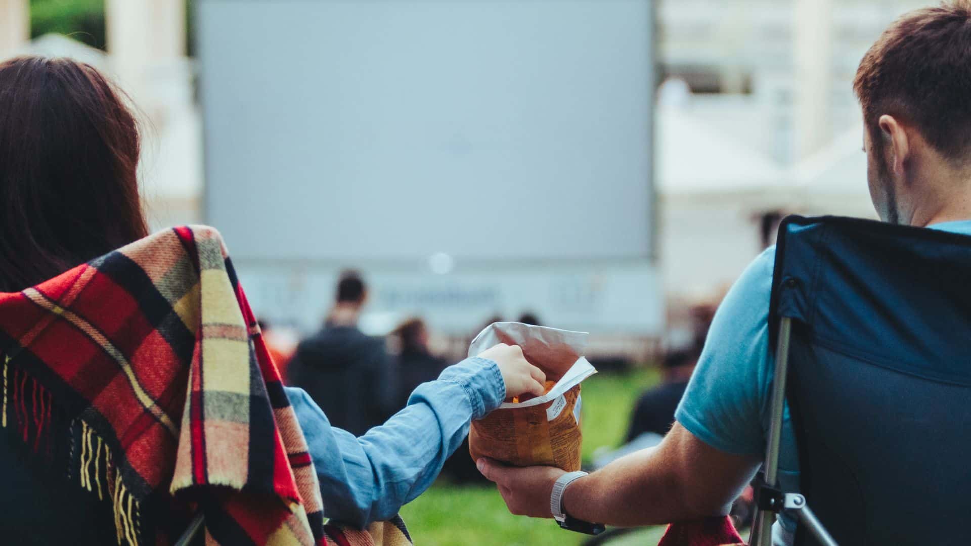 A couple share snacks in front of a cinema screen in a city park.